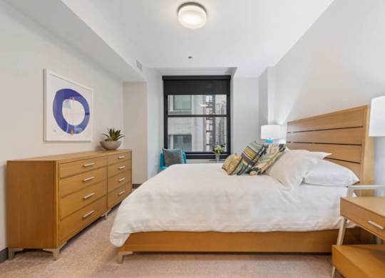 A bedroom with a large bed, a wooden dresser, and a window with a view of a cityscape.