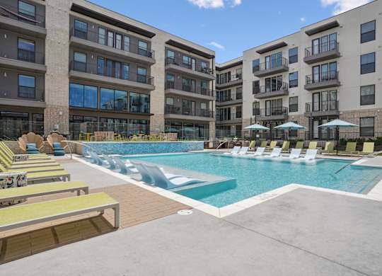 A swimming pool surrounded by sun loungers and umbrellas in front of apartment buildings.