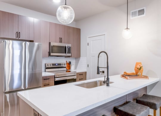 2-Bedroom Apartments in Raymore, MO - The Venue Townhomes - Designer Kitchen with Island, Pantry Stainless Steel Samsung Appliances, Wood-Style Cabinetry, White Tile Backsplash, White Quartz Countertops, Bar-Stool Seating, and Pendant Lights.