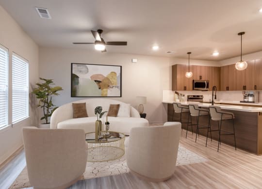 Apartments for Rent in Raymore, MO - The Venue Townhomes - Furnished Living Room with Designer Luxury Vinyl Tile Flooring, Area Rug, Sofa, Armchairs, Round Glass-Top Coffee Table, Side Table with Lamp, Houseplant, Ceiling Fan, Large Window, and Open-Concept Layout with View of Kitchen with Bar-Style Chairs.