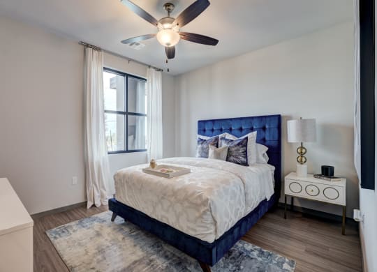 Cozy bedroom here at 35 Degrees North Apartment Homes with a blue tufted bed, white bedding, and decorative pillows. A tray sits on the bed. A lamp and decor adorn the nightstand. Soft, natural light filters through the window, creating a serene ambiance.