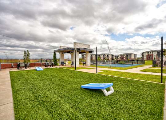 Outdoor recreational area here at 35 Degrees North Apartment Homes with a cornhole game on green grass, white pergola with seating, tennis court in background, overcast sky and string lights.