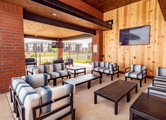 Covered patio here at 35 Degrees North Apartment Homes with striped cushioned seating, wooden ceiling, and brick walls. A large TV is mounted on the wooden wall. Apartments are visible outside.