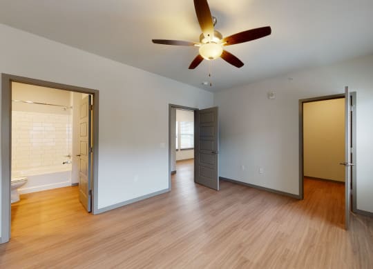 Empty room here at 35 Degrees North Apartment Homes with wood floors, gray doors, and a ceiling fan. Open doors reveal a bright bathroom with white tiles and a window-lit adjacent room.
