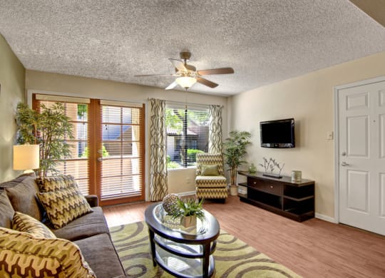 Modern living room here at Desert Horizon Apartments with beige walls, a gray sofa with patterned cushions, glass coffee table, and wall-mounted TV. Natural light from large windows.