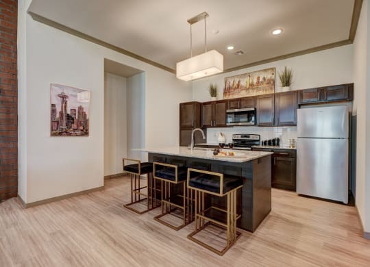 Modern kitchen here at 35 Degrees North Apartment Homes with dark wood cabinets and a granite island. Four sleek stools surround the island, with wine glasses and a snack on top. Warm lighting and art enhance the cozy ambiance.