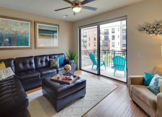 Modern living room here at Inspire Apartment Homes with a black leather sectional, colorful cushions, large abstract art, and sliding doors leading to a balcony with blue chairs. Cozy ambiance.