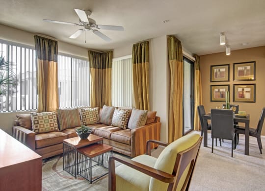 Modern living room here at Natura Villas Apartments with a brown leather sectional sofa, geometric pillows, and a wooden coffee table. Dining area features a black table set for four, with abstract wall art. Warm, neutral tones create a cozy ambiance.
