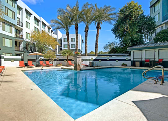 Outdoor pool here at Acclaim Apartments surrounded by modern apartment buildings and palm trees. Red lounge chairs and umbrellas beside the pool create a relaxing atmosphere.