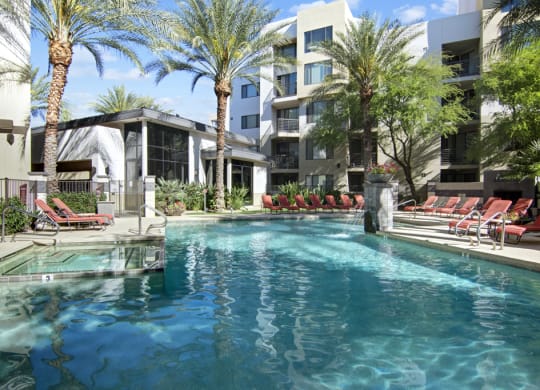 Outdoor pool area here at Acclaim Apartments with clear blue water surrounded by red lounge chairs and palm trees, adjacent to modern white buildings under a bright, sunny sky.