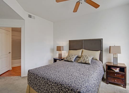 Cozy bedroom here at Acclaim Apartments with a queen bed, dark patterned bedspread, two wooden nightstands with lamps, and a ceiling fan. Doorway to a bathroom is visible.