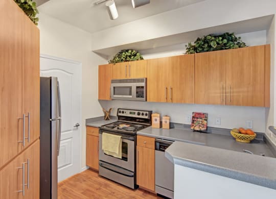 Modern kitchen here at Acclaim Apartments with wood cabinets, stainless steel appliances, and a gray countertop. Decor includes greenery, a recipe book, and a fruit bowl. Cozy and organized ambiance.