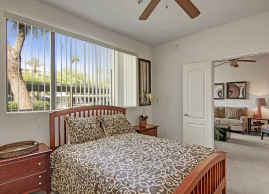 Bedroom here at Acclaim Apartments with a patterned bedspread, wooden nightstand, and potted plant. Open door reveals a cozy living room with a sofa and two paintings. Bright, calming atmosphere.