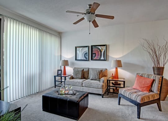 Stylish living room here at Acclaim Apartments with a gray couch, striped chair, and glass-topped black ottoman. Modern art, orange accents, tall vase, sunlight through blinds.