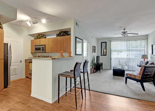 Modern open-plan living room and kitchen here at Acclaim Apartments with wooden flooring, light walls, and neutral decor. A countertop with bar stools divides the spaces. A ceiling fan and large window with vertical blinds add to the bright, airy feel.