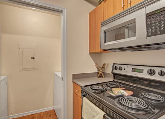 A small kitchen here at Acclaim Apartments with a black electric stove, stainless steel microwave, and wooden cabinets. Adjacent is a laundry alcove with a washing machine, evoking a compact, efficient space.