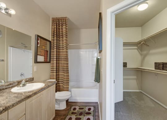 Modern bathroom here at Natura Villas Apartments with a granite countertop, a sink, and a large mirror on the left. A bathtub with patterned shower curtain is in the center. An open door on the right leads to a walk-in closet with shelving. Warm lighting creates a cozy atmosphere.
