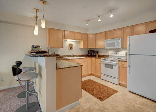 A spacious kitchen here at Admirals Cove with light brown cabinets, granite counter tops, bar type seating, and a ceramic sink. The room has a warm tone.
