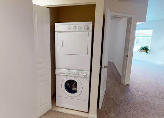 A hallway laundry closet at Admirals Cove featuring a stacked washer and dryer set behind a partially open door. Soft neutral carpet and nearby bedroom and living room entrances create an easy flow through the home, offering convenient in-unit laundry access.