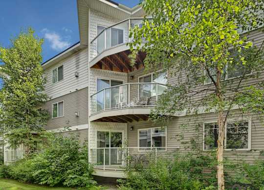 A three-story exterior view of Admirals Cove showcasing private balconies with glass railings surrounded by tall trees and green landscaping. Light siding and shingle detailing give the building a clean, modern look. Outdoor seating on some balconies highlights a relaxing residential setting.