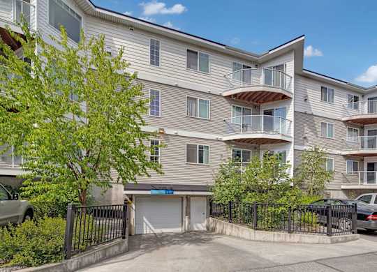 A multi-story exterior view of Admirals Cove featuring light siding, private balconies with glass railings, and landscaped greenery along the front. Parking is visible near the entrance, and a gated driveway leads to underground garage access beneath the building, showcasing convenient resident amenities.