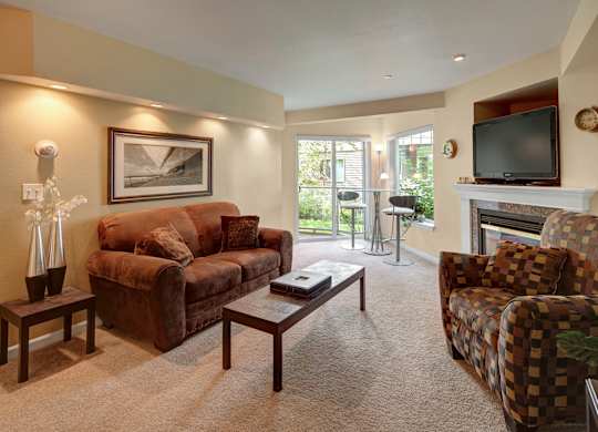 A cozy living room here at Admirals Cove with a light brown carpet and soft lighting. There is a brown couch, and a brown, checkered patterned sofa chair. There is a two-person table near a sliding glass door that leads you outside.