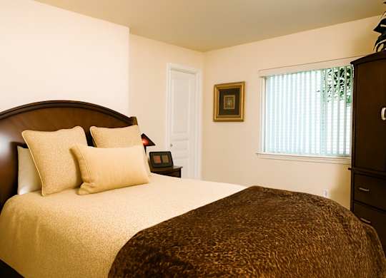 A cozy bedroom at Admirals Cove with warm cream walls and soft natural light filtering through window blinds. The room features a neatly made bed with plush pillows and a dark wood headboard, complemented by matching furniture pieces and simple wall art for a comfortable, inviting feel.