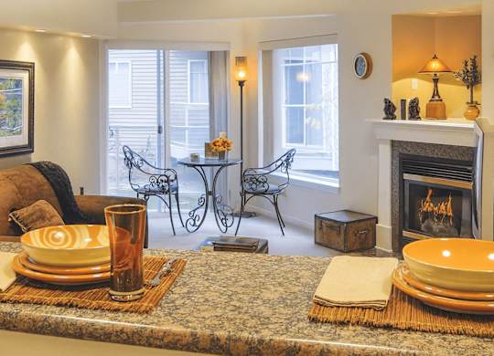 A warm living area at Admirals Cove with a cozy seating space and a fireplace along the wall. A small dining nook with two decorative chairs sits beside large windows and a sliding door leading to the patio. The granite countertop in the foreground is set with colorful dishes, giving the space a welcoming feel.