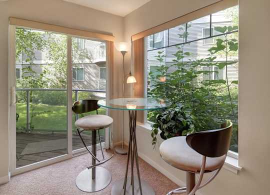 A bright dining nook at Admirals Cove featuring a round glass table with two modern bar-height chairs. Large windows and a sliding glass door overlook green landscaping, filling the space with natural light. Soft carpeting and corner lighting create a relaxed spot for meals or morning coffee.