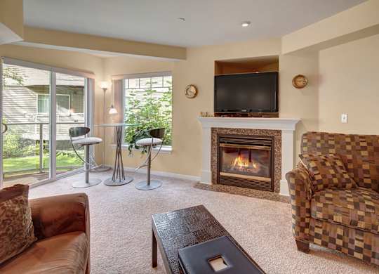 A bright living room at Admirals Cove with soft carpet flooring and a cozy seating area. A fireplace with a TV above serves as the focal point, while large windows and a sliding door bring in natural light. A small glass-top dining set sits beside greenery just outside, creating a relaxing indoor-outdoor feel.