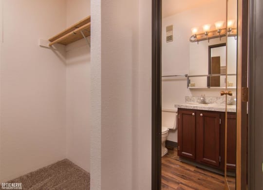 Compact bathroom here at Outlook Apartments with wooden cabinets, marble countertop, and lit wall mirror seen from a doorway. Adjacent closet with a wooden shelf. Warm and clean ambiance.