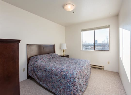 Simple bedroom here at Outlook Apartments with a paisley-patterned bed, wooden headboard, and nightstand with lamp. Window shows cityscape, creating a serene atmosphere.
