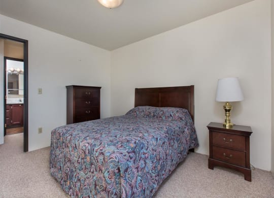 A cozy bedroom here at Outlook Apartments features a bed with a colorful paisley bedspread, a wooden nightstand with a brass lamp, a tall dresser, and a doorway leading to a bathroom.