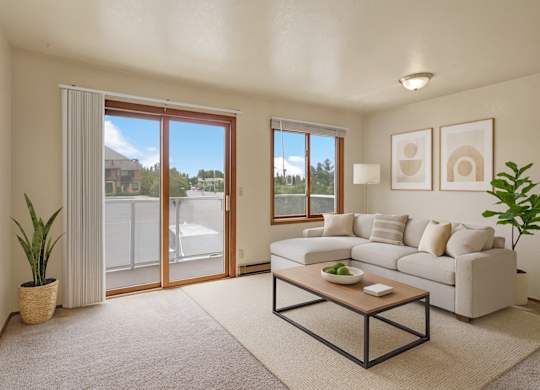 Bright living room at Arbor Pointe with a large beige sectional sofa, wooden coffee table, indoor plants, and sliding glass door leading to a balcony with views of nearby buildings and trees.