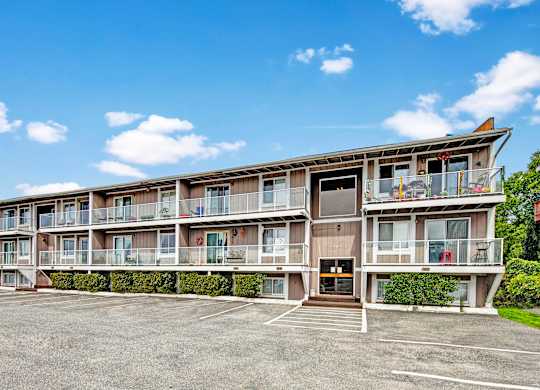 Exterior view of Arbor Pointe apartment building with balconies, large windows, and a parking lot in front under a blue sky with scattered clouds.