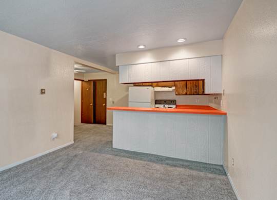 Another angle of an unfurnished Arbor Pointe living area with soft gray carpet and a small breakfast bar leading into a compact kitchen with bright orange countertops, white appliances, and wood upper cabinets