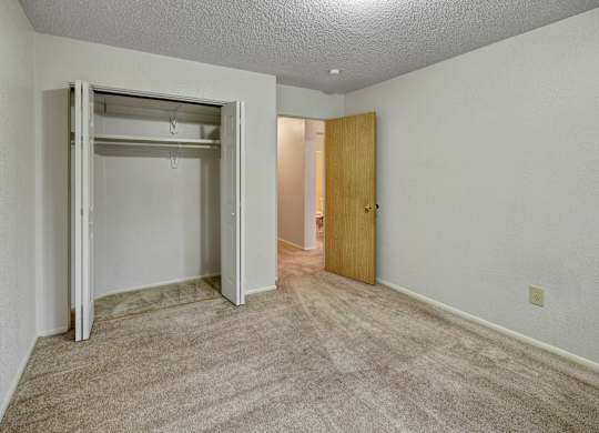 A bright bedroom at Arctic Sun Apartments with plush beige carpet and neutral walls. An open closet with double doors offers storage space. A doorway leads to the hallway and bathroom, and a ceiling light adds soft illumination, creating a comfortable and versatile room layout.
