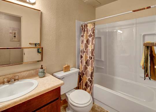 A warm bathroom at Arctic Sun Apartments with a tub and shower combo and a patterned curtain. The space includes a vanity with a sink, a wide mirror, and storage below, along with a toilet. Soft lighting and neutral tones create a comfortable, practical layout.