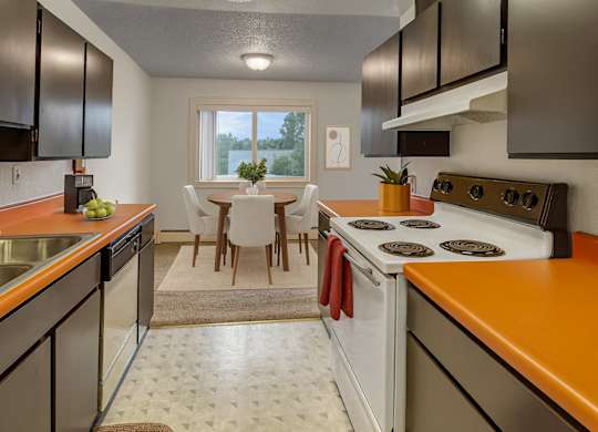 A bright kitchen at Arctic Sun Apartments featuring dark cabinets, orange countertops, a stainless sink, dishwasher, and stovetop. The space opens to a dining area with a round table, four upholstered chairs, and a window overlooking trees, creating a warm and inviting eating area.