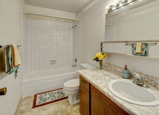 A clean bathroom at Arctic Sun Apartments featuring a bathtub and shower combo with tiled walls, a toilet, and a long marble-style countertop with a sink. A wide mirror with vanity lighting brightens the space, and towel racks and decor add color and warmth.