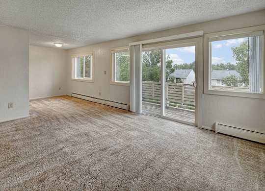 A bright living space at Arctic Sun Apartments with plush beige carpet, neutral walls, and large windows. A sliding glass door opens to a private balcony with neighborhood views, bringing in abundant natural light. The open layout offers flexible room for seating and dining arrangements.
