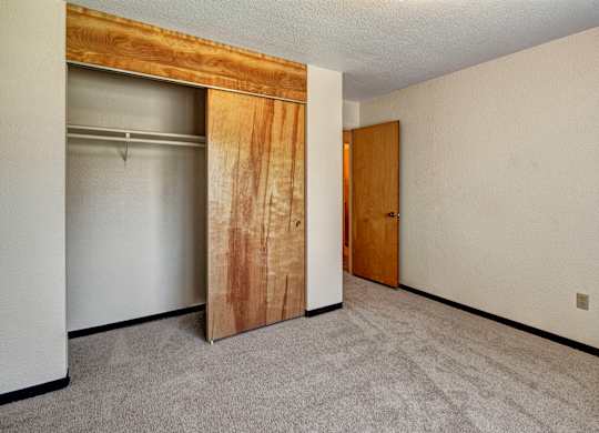 A clean bedroom at Bay Arms Apartments with plush carpet and neutral walls. A large closet with sliding wood doors provides storage space, and a doorway leads to the hallway. The room’s simple layout offers plenty of flexibility for bedroom furnishings.