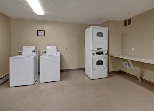 A clean laundry room at Bay Arms Apartments featuring two top-loading washing machines and a stacked dryer set. A long folding counter provides workspace, and neutral walls and flooring create a bright, practical environment for everyday laundry needs.