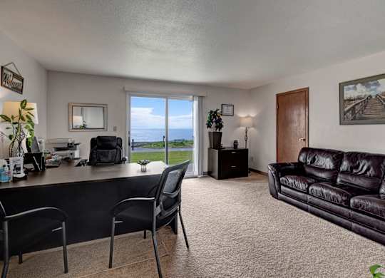 A welcoming leasing office at Bay Arms Apartments with a desk and chairs, a leather sofa, and décor. A large sliding glass door offers a stunning view of the water and grassy outdoor space, filling the room with natural light. Soft carpet and neutral walls create a comfortable environment for residents and guests.