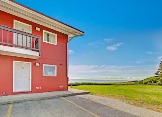 A bright exterior view of Bay Arms Apartments showing part of a red two-story building with white trim and private entrances. A wide grassy lawn stretches out beside the parking area, leading to stunning ocean views and a clear blue sky in the distance.