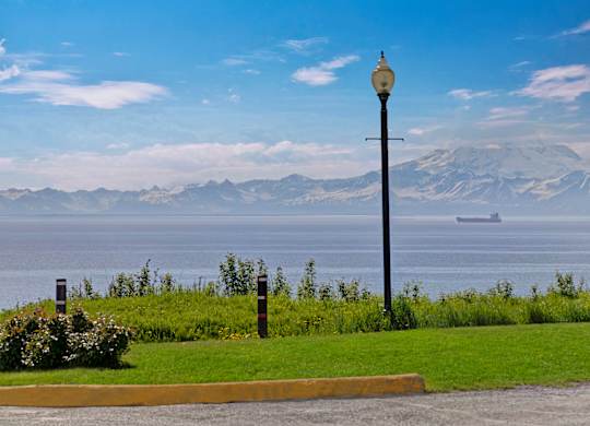 A breathtaking oceanfront view at Bay Arms Apartments with lush green lawn, a lamp post, and distant mountains rising over the water. A cargo ship is visible on the horizon, and the clear blue sky adds to the peaceful coastal setting.