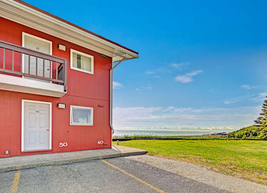 A bright exterior view of Bay Arms Apartments showing part of a red two-story building with white trim and private entrances. A wide grassy lawn stretches out beside the parking area, leading to stunning ocean views and a clear blue sky in the distance.