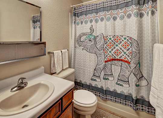 A cozy bathroom at Bay Arms Apartments with a vanity and sink, a wide mirror, and a shower and bathtub combo. A patterned curtain with an elephant design adds color and personality. The room includes a toilet, towel rack, and neutral walls, creating a clean and welcoming space.