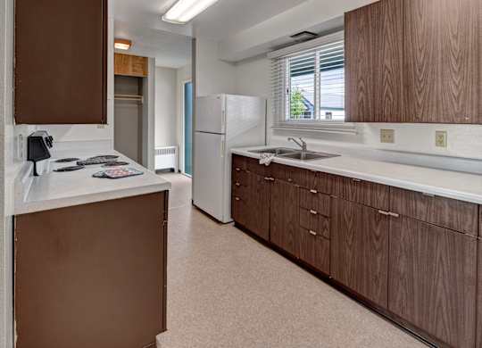 A spacious kitchen at Bay Arms Apartments featuring wood cabinets, long countertops, a double sink, and a stovetop. A large refrigerator sits near a bright window with blinds that bring in natural light. Neutral flooring and ample storage make the space functional and convenient.