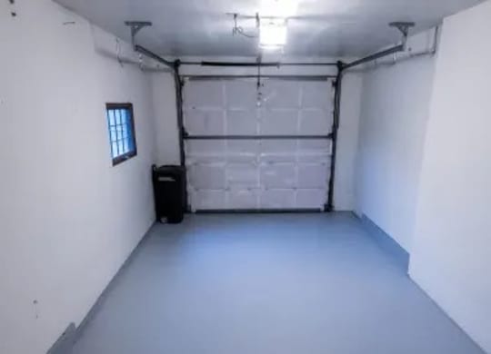 A clean Birchwood garage with freshly painted pale blue floors and bright white walls, offering a tidy single-car space beneath overhead lighting. A small window brings natural light into the room, while a black utility bin sits neatly near the closed garage door.